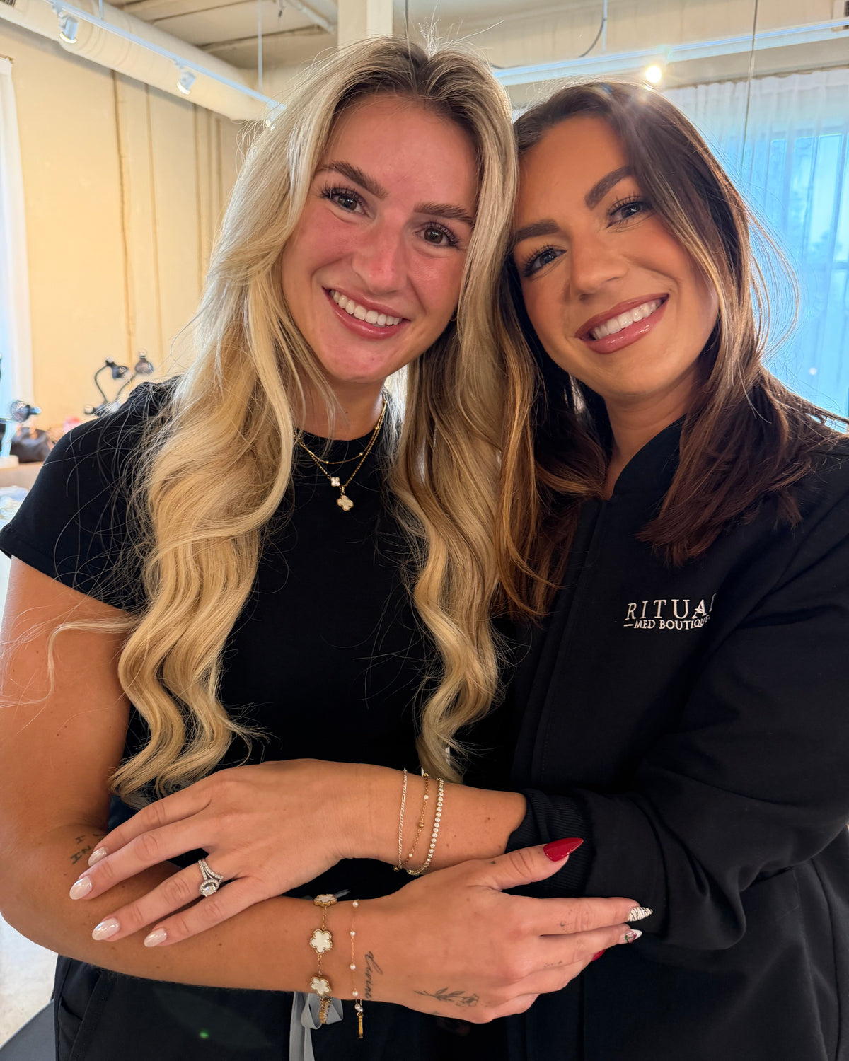 Two women smiling and posing together indoors. They are wearing their Everlasting Permanent Jewelry 